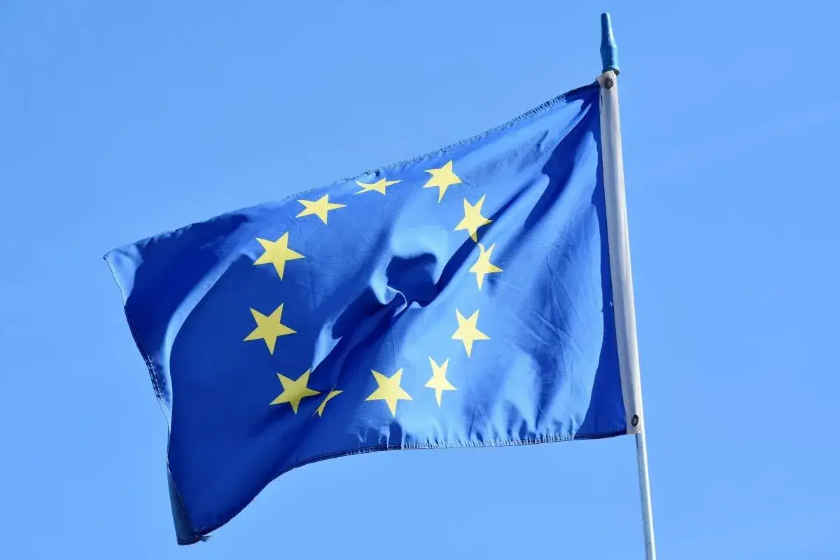Błękitna flaga z dwunastoma złotymi gwiazdami tworzącymi okrąg, symbolizująca godło Unii Europejskiej, powiewa na maszcie na tle błękitnego nieba.