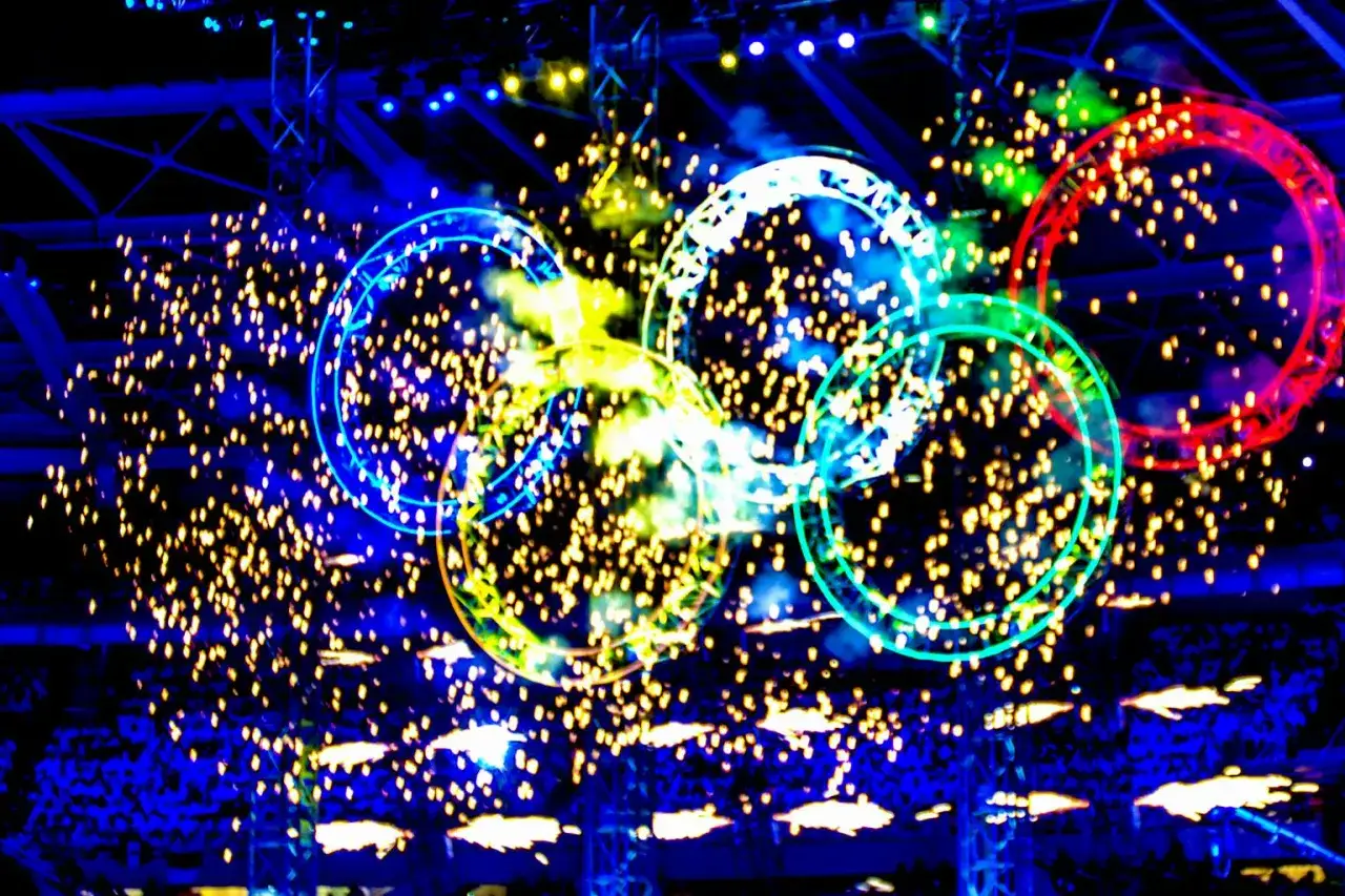 Kolorowe kręgi symbolizujące flagę olimpijską unoszą się wśród złotych iskier fajerwerków.