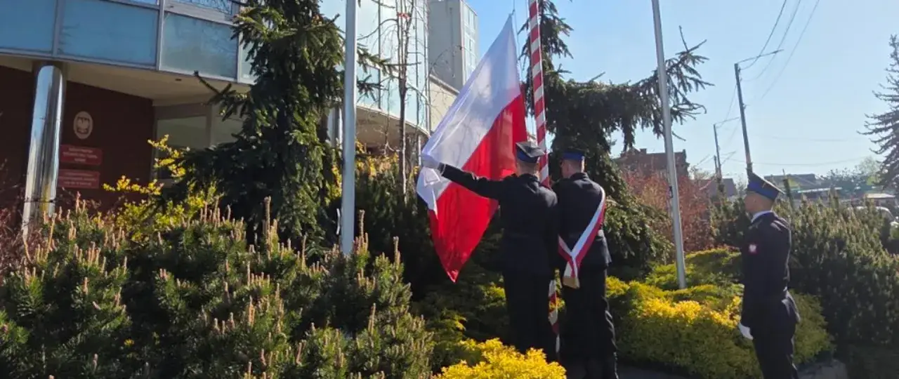 Uroczyste podniesienie flagi na maszcie przez dwóch żołnierzy w mundurach. Biało-czerwona flaga powiewa na wietrze.
