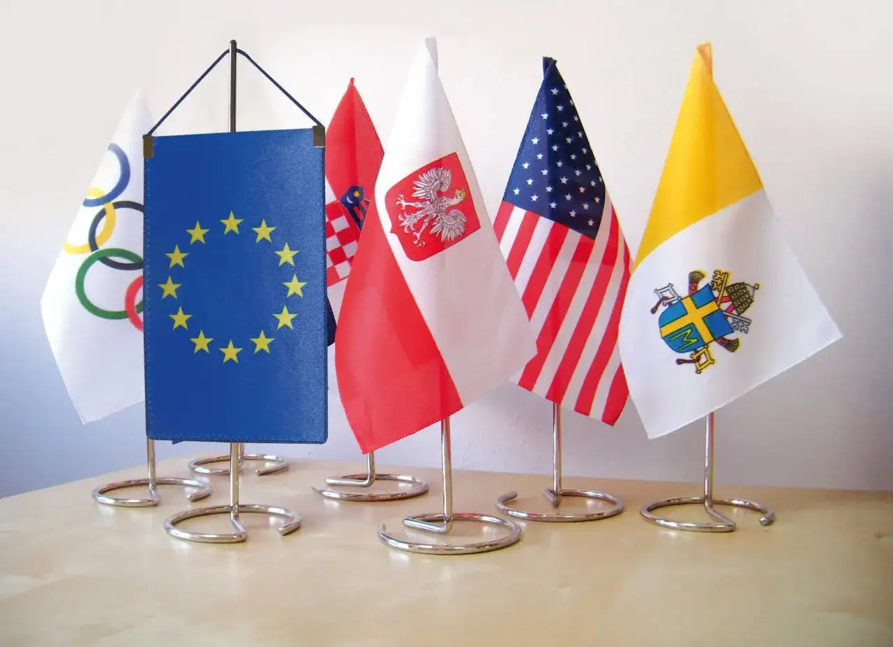Na stole stoi kilka małych flag: olimpijska, Unii Europejskiej, Polski, USA i Watykanu. Każda chorągiewka na metalowym stojaku.