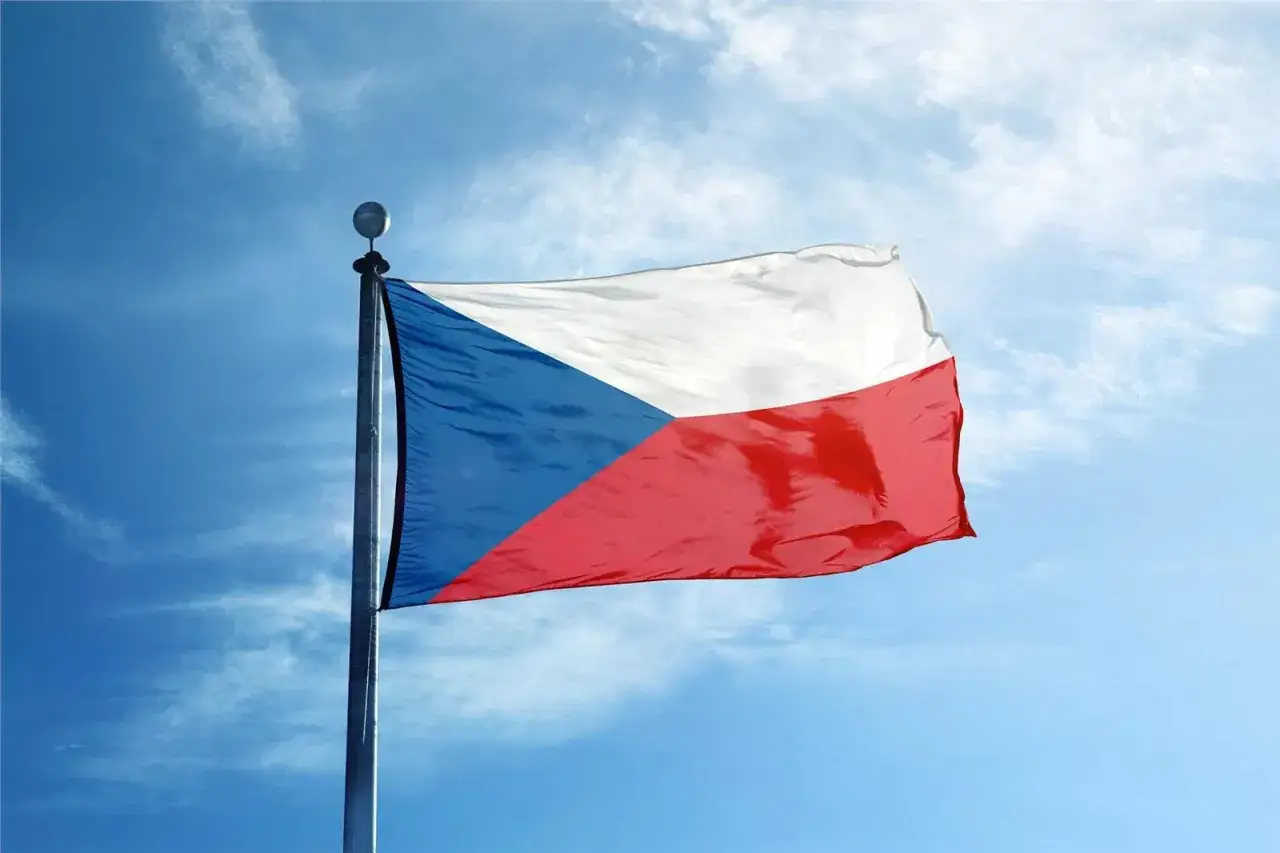 Flaga Czech powiewa na maszcie na tle błękitnego nieba z chmurami. Biały, niebieski i czerwony pas tworzą charakterystyczny wzór.
