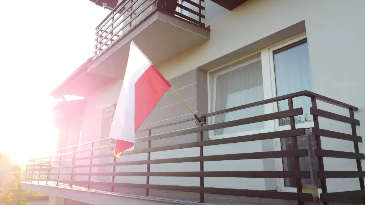 Flaga Polski powiewa pionowo na balkonie, ozdabiając dom w promieniach zachodzącego słońca.
