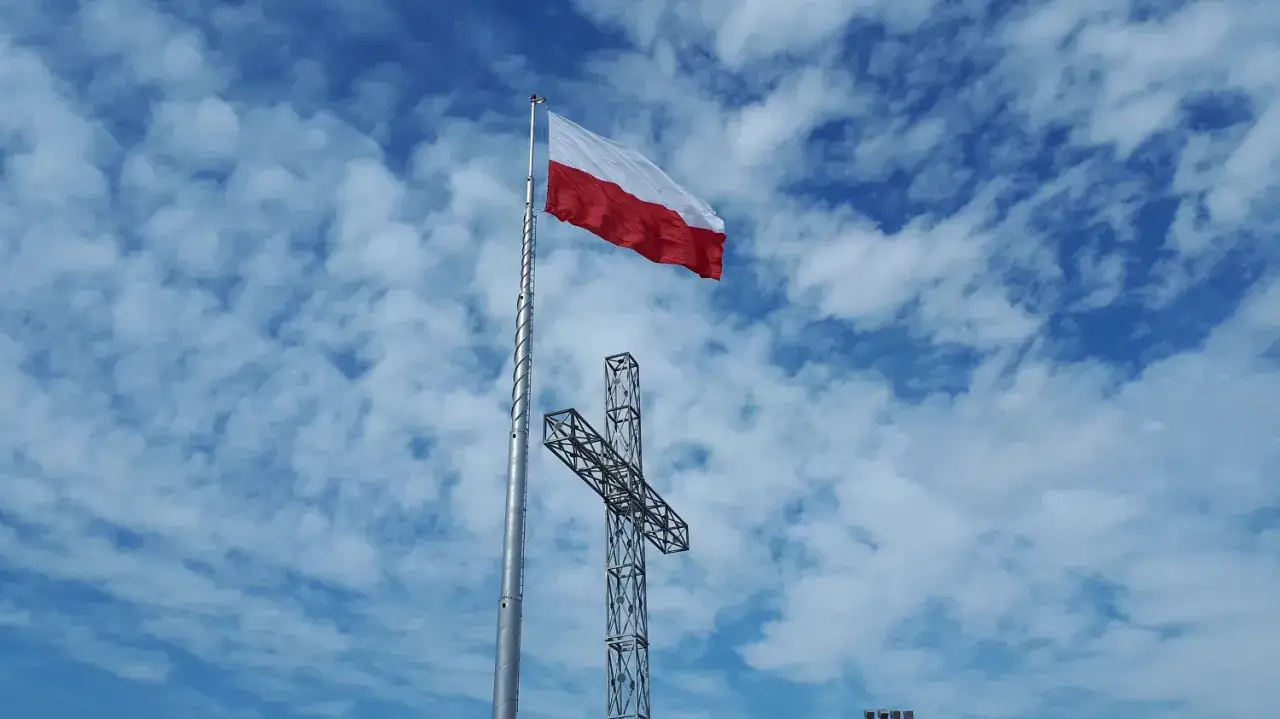 Biało-czerwona flaga państwowa powiewa na maszcie obok metalowego krzyża na tle błękitnego nieba z chmurami.