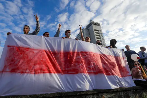 Ludzie trzymają biało-czerwoną flagę, symbol wolności i nadziei. Na niebie chmury, w tle budynek.
