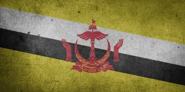 Flaga Brunei z godłem w kolorze czerwonym, na tle ukośnych pasów: żółtego, białego i czarnego.