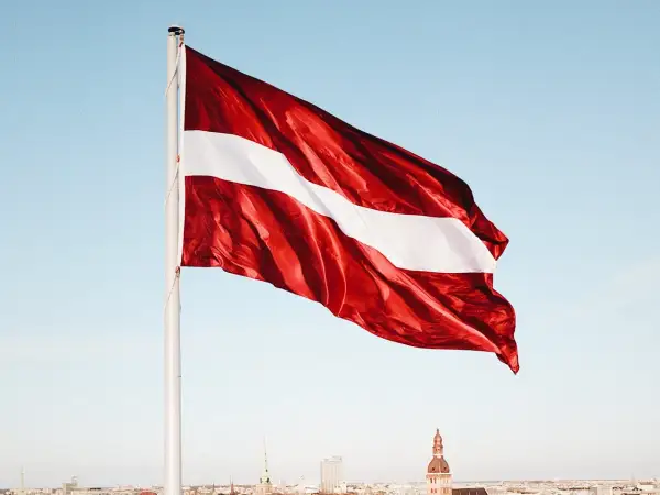 Flaga Łotwy: ciemnoczerwona z białą wstęgą pośrodku, na tle błękitnego nieba i miasta.