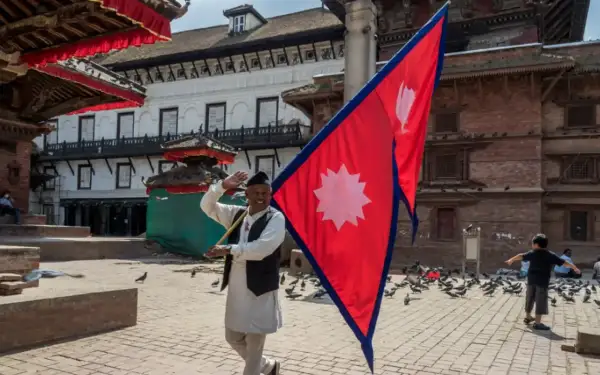 Mężczyzna macha flagą Nepalu na placu pełnym gołębi. W tle tradycyjna architektura.