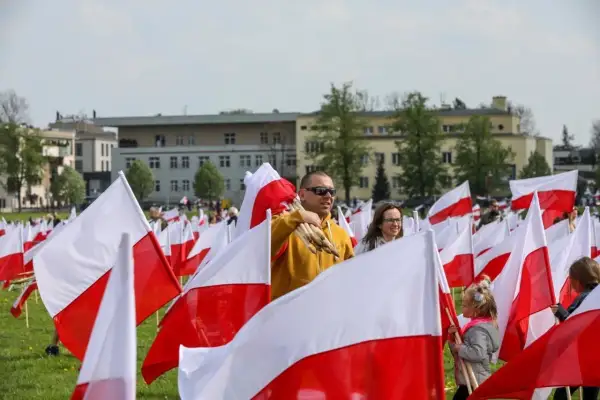 Ludzie z biało-czerwonymi flagami, wśród nich mężczyzna z pęczkiem flag. W tle budynki i drzewa. Flaga Torunia dumnie powiewa.