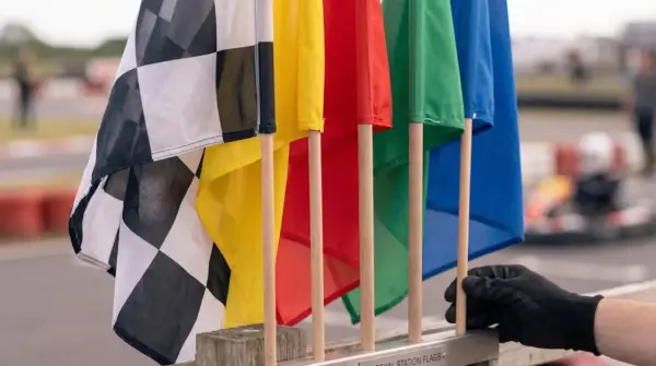 Flaga wyścigowa w kratkę obok kolorowych flag: żółtej, czerwonej, zielonej i niebieskiej. W tle widać tor wyścigowy i gokarta.